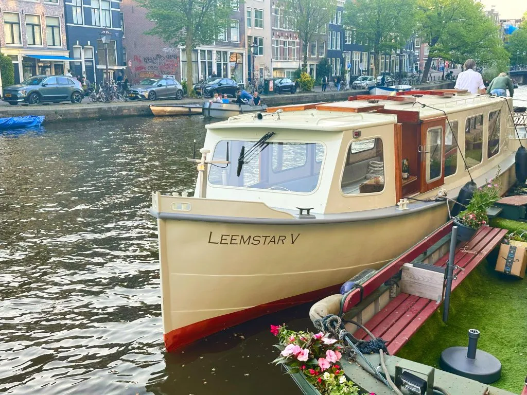 Leemstar V salonboot, afgemeerd aan een groene kade met bloemen, met de Amsterdamse grachtenpanden op de achtergrond.