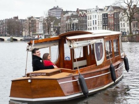 Couple on a wooden boat in an Amsterdam canal with buildings in the background.