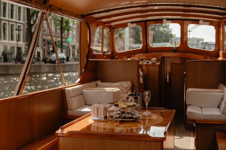 Interior of a luxury boat with wooden furnishings, table, and cushioned seats on a canal.