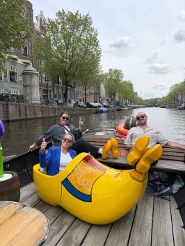 Three people giving thumbs up in a boat, one sitting in a giant yellow clog, on a canal.