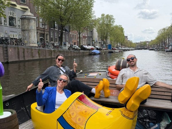 Three people giving thumbs up in a boat, one sitting in a giant yellow clog, on a canal.