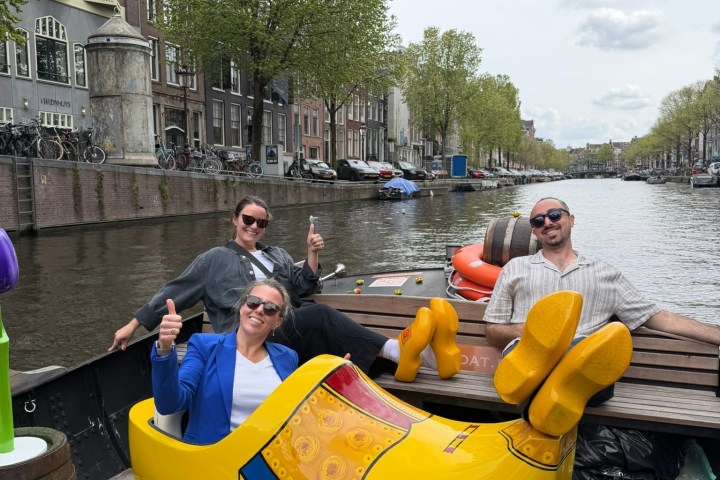 Three people giving thumbs up in a boat, one sitting in a giant yellow clog, on a canal.