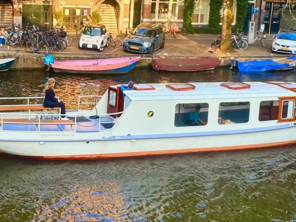 White canal boat with people seated, passing by parked bicycles and cars.