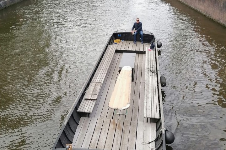 Man steering a large, empty boat on a canal with lined trees and parked cars.