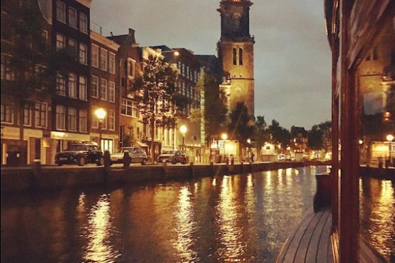 Canal at dusk with lit streetlights and tall clock tower reflecting in water.