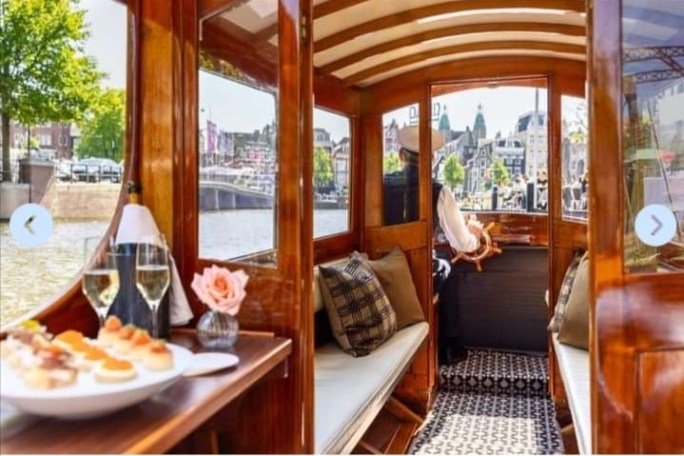 Inside a wooden boat with a meal and drinks, man steering, view of a canal with buildings and trees.