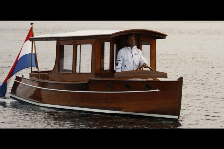 Wooden boat with a red, white, and blue flag on calm water, piloted by a person in a white jacket.