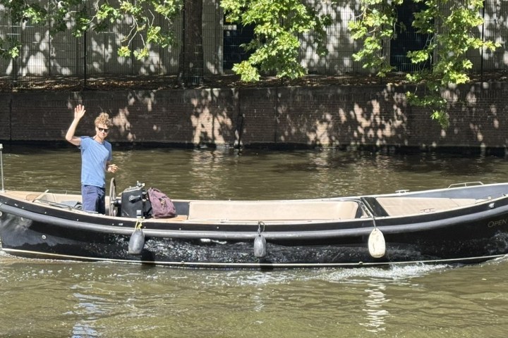 Man waving on a black boat sailing on a river with trees in the background.