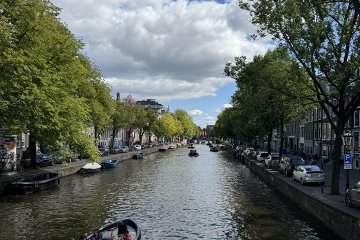 Boats on a canal with trees on both sides, flowers in foreground, under a cloudy blue sky.