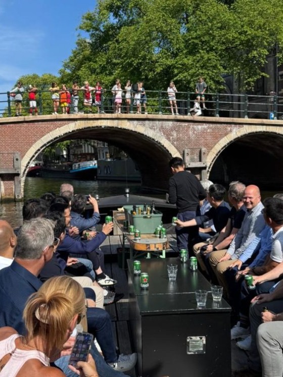 People on a boat tour in a canal, with a bridge and onlookers in the background, surrounded by trees.