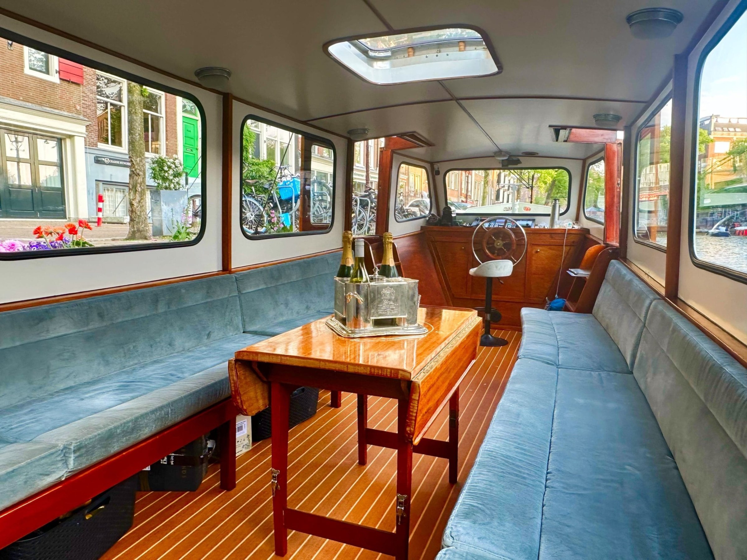 Luxury boat interior with blue seating, wooden floor, table, champagne, and steering wheel.