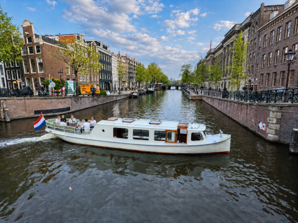Boat on a canal in a city with buildings and trees lining the sides, under a partly cloudy sky.