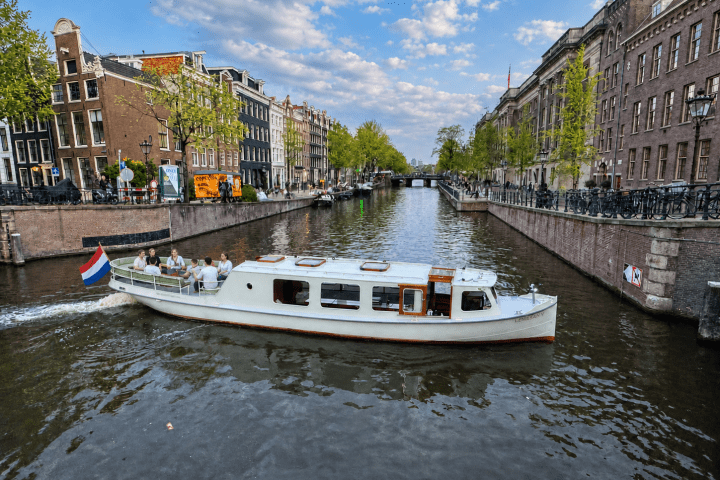 Boat on a canal in a city with buildings and trees lining the sides, under a partly cloudy sky.
