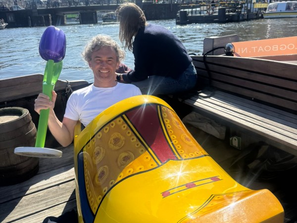 Person sitting in giant yellow clog holding inflatable flower by waterfront.