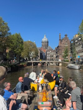 People on a boat tour in a sunny Amsterdam canal, with historic buildings and trees lining the water.