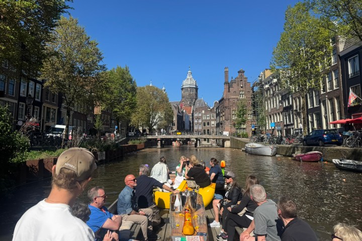 People on a boat tour in a sunny Amsterdam canal, with historic buildings and trees lining the water.