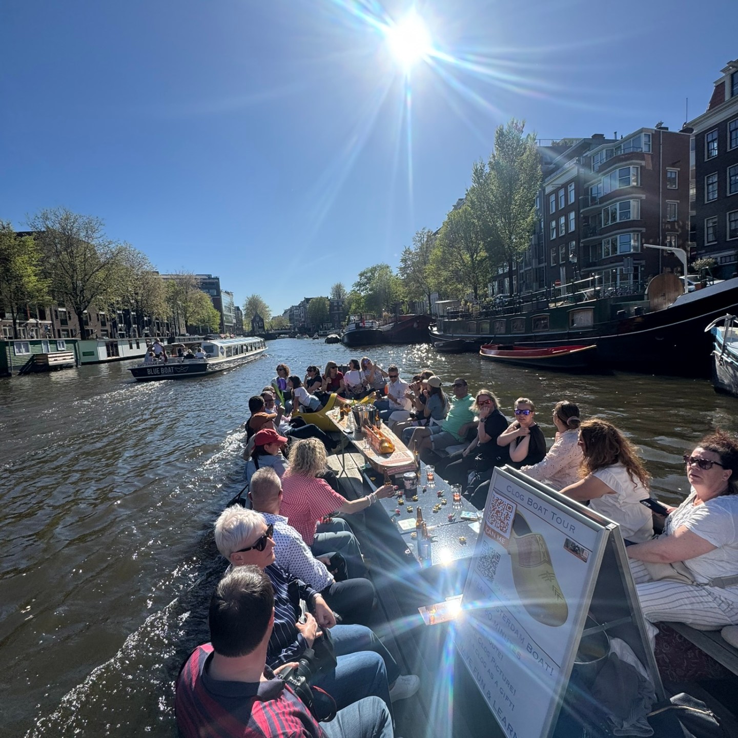 People on a boat cruise through a sunny canal in a city with trees and buildings.