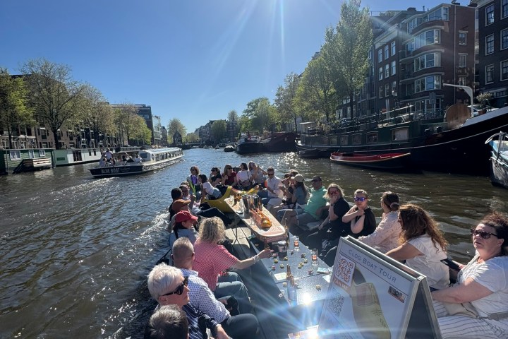 People on a boat cruise through a sunny canal in a city with trees and buildings.