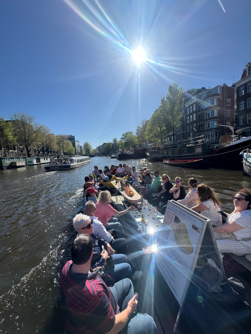 People on a boat cruise through a sunny canal in a city with trees and buildings.